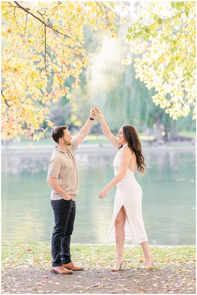 The man twirls the woman under his raised arm near the pond in Boston Public Garden. The woman’s white dress flows with the motion, while colorful autumn leaves hang above them.