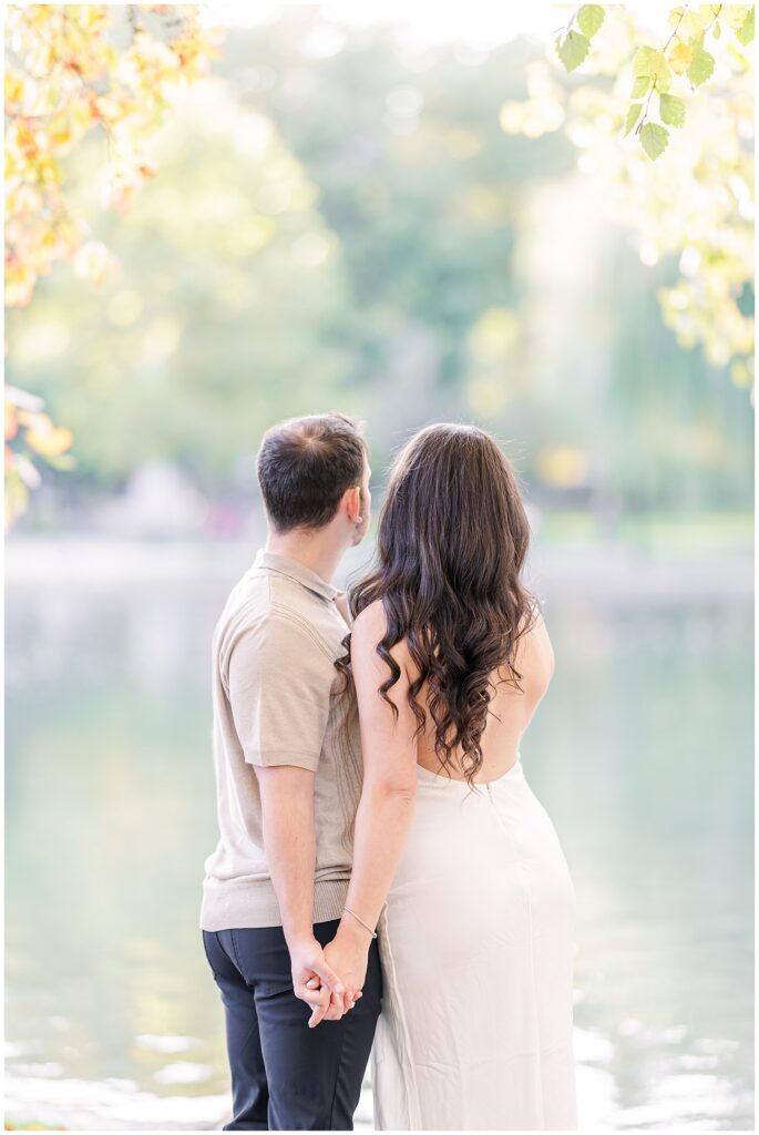 The couple stands together, facing away from the camera, holding hands near the pond at Boston Public Garden. The woman’s long, wavy hair flows over her white dress as they look out toward the peaceful scenery.