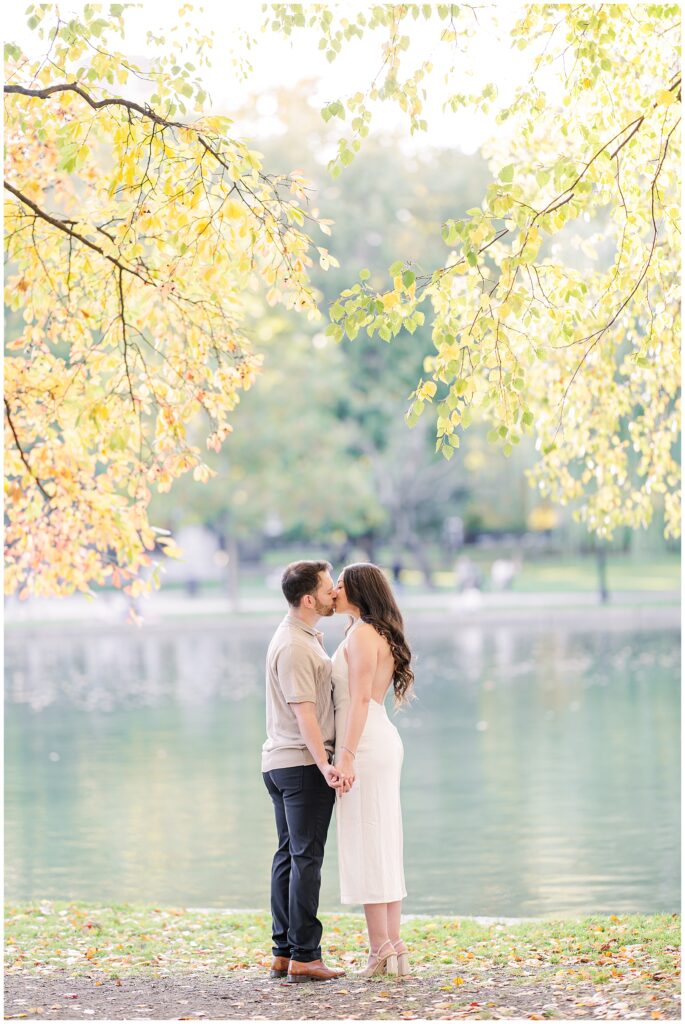 A couple holds hands and kisses by the pond at Boston Public Garden, framed by golden autumn leaves. The serene water and vibrant foliage create a romantic setting perfect for Boston engagement pictures.