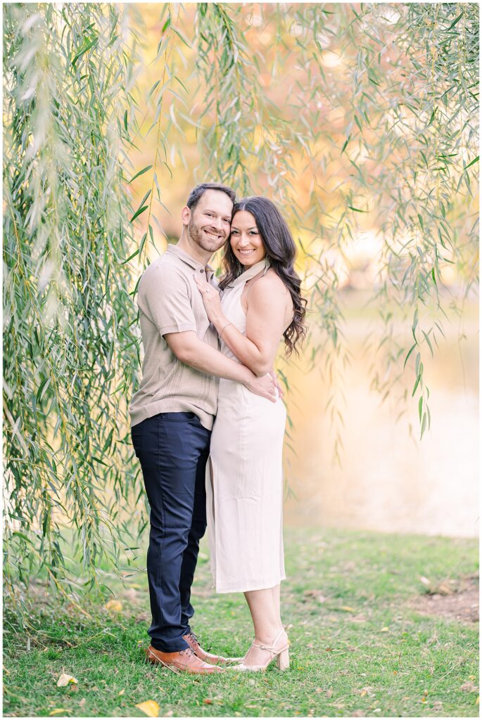 The couple stands together under hanging willow branches at Boston Public Garden. The man wears a beige shirt, and the woman is in a white dress, both smiling warmly against the soft fall tones.