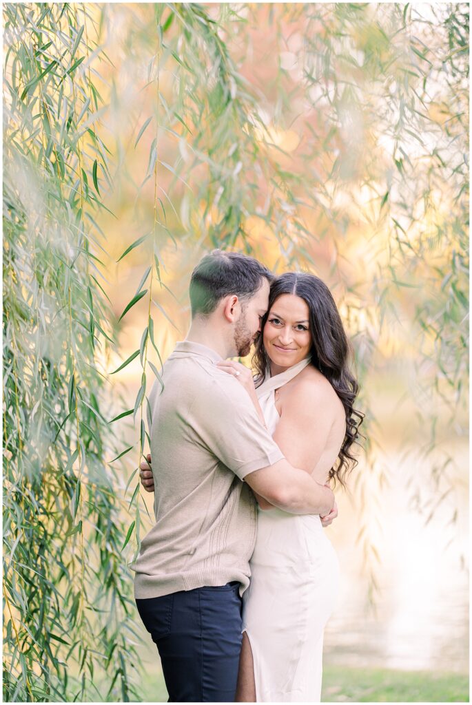 The man embraces the woman as they stand close under willow branches at Boston Public Garden. The woman smiles gently at the camera while the man leans his head toward her, surrounded by golden hues.