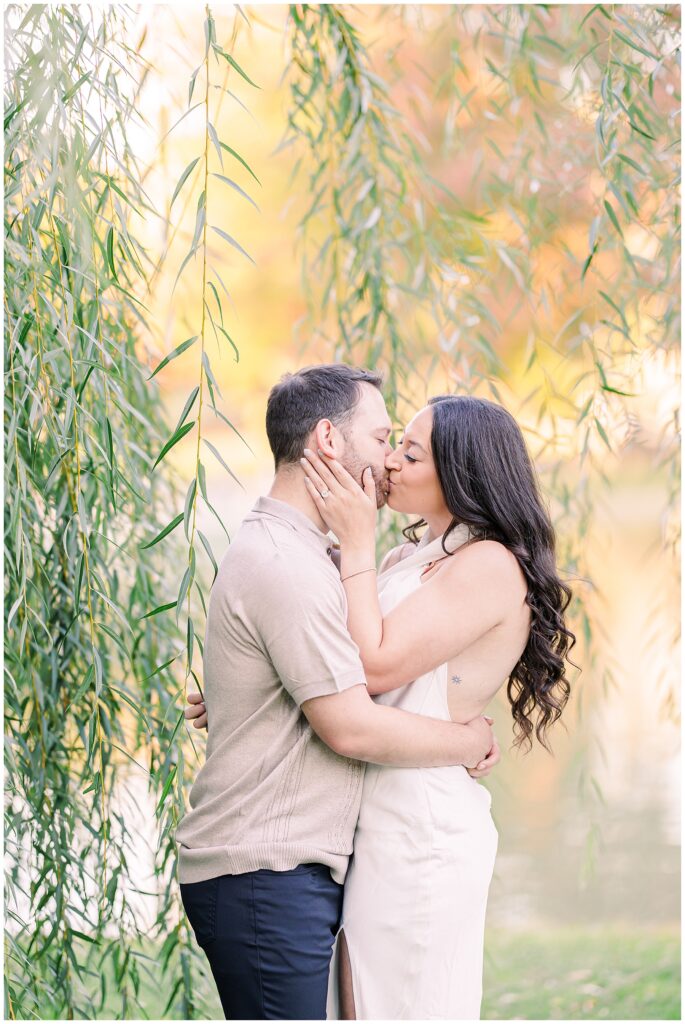 The couple shares a kiss under the willow branches at Boston Public Garden. The woman cups the man’s face with one hand, her engagement ring visible as soft fall colors frame the moment.