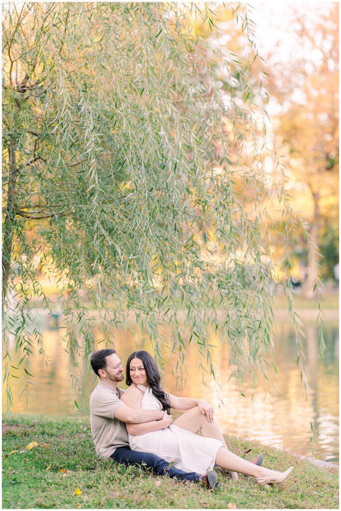 The couple sits on the grass near the pond in Boston Public Garden, surrounded by hanging willow branches. The man wraps his arms around the woman as she smiles, with autumn tones reflected in the water.