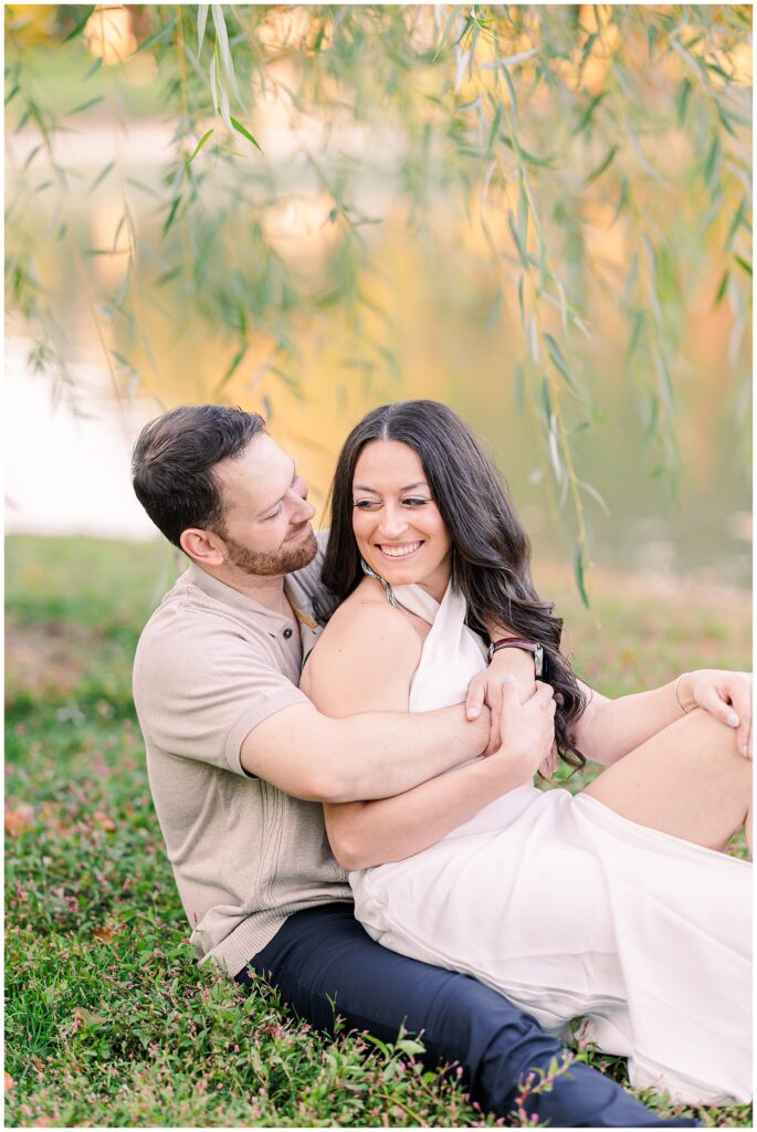 The couple sits closely on the grass at Boston Public Garden. The woman leans back into the man’s arms, both smiling warmly, with soft willow branches and the pond in the background.