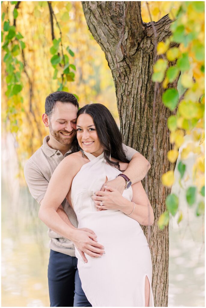 The couple poses under a tree at Boston Public Garden, with golden leaves framing them. The man has his hand in his pocket while the woman lifts one leg playfully, creating a cheerful moment.