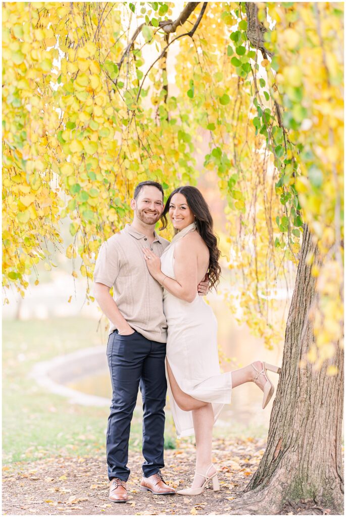 The couple leans against a tree at Boston Public Garden, smiling as the man wraps his arms around the woman. The golden leaves and pond add a soft, romantic atmosphere to this Boston engagement picture.