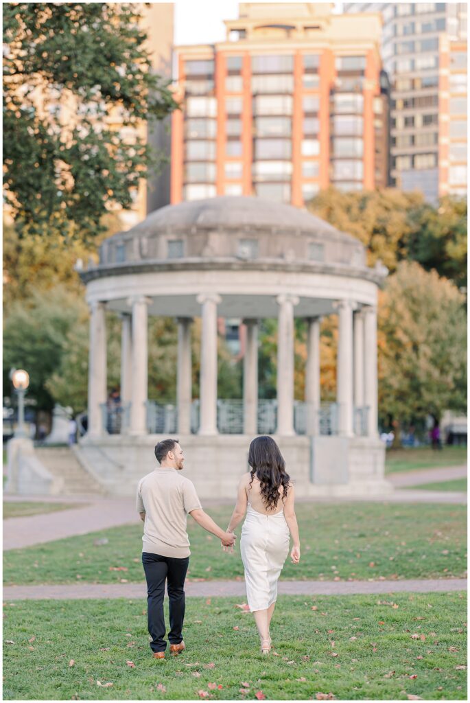 A couple walks hand-in-hand through Boston Common, heading toward the iconic Parkman Bandstand. The man looks at the woman, who wears a white dress, as the lush greenery and city buildings form the backdrop. A picturesque moment for Boston engagement pictures.