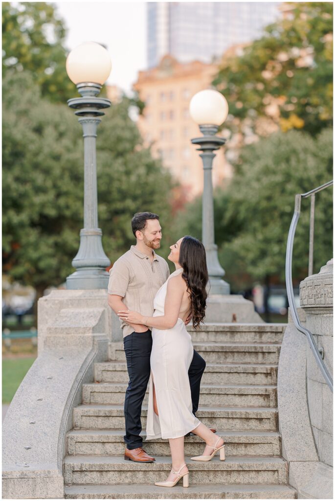 The couple stands on the steps of the Parkman Bandstand in Boston Common, gazing at each other with soft smiles. The vintage-style lampposts and trees frame this romantic Boston engagement picture.