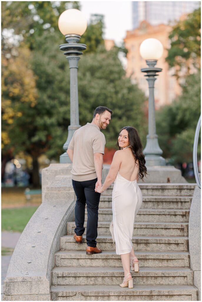 The couple walks up the steps of the Parkman Bandstand in Boston Common, holding hands. The woman turns to smile at the camera, her flowing white dress adding elegance to this engagement scene.