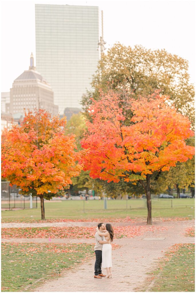 The couple embraces on a path in Boston Common, surrounded by vivid red and orange autumn trees. The Boston skyline rises in the background, blending urban and natural beauty, ideal for Boston engagement pictures.