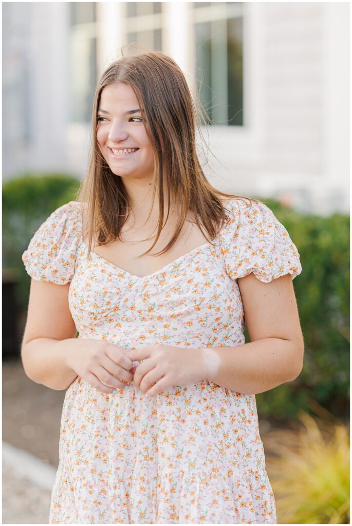 A high school senior girl stands outdoors, smiling and looking to her left. She has long, straight brown hair and is wearing a white floral dress with small orange and green flowers. The background is softly blurred with greenery and part of a light-colored building.