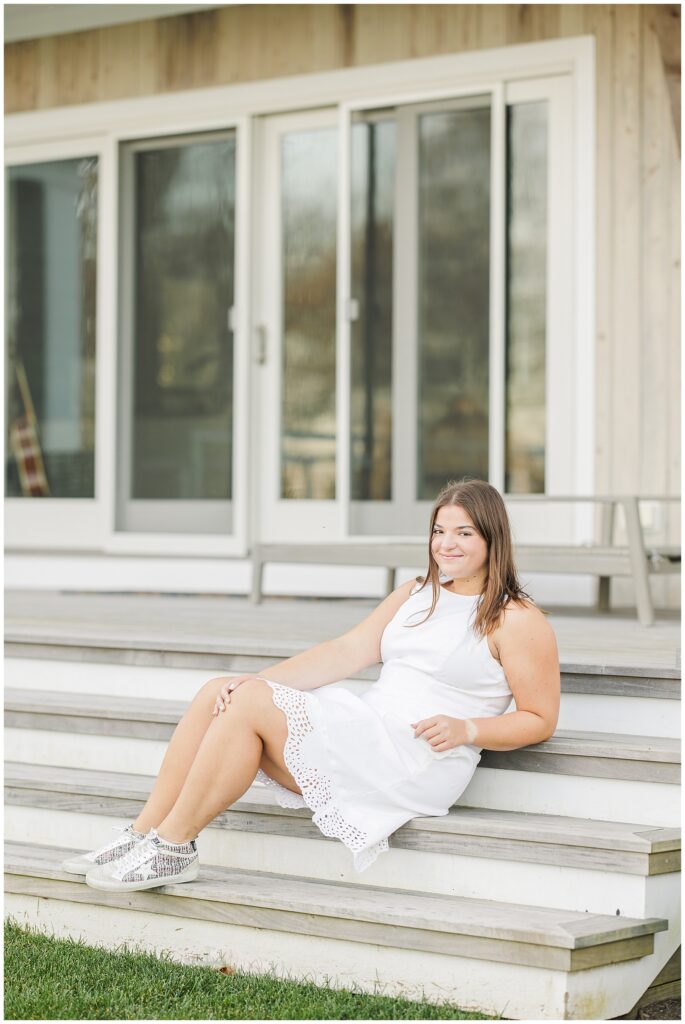 The senior girl is sitting on the steps of a porch in front of a light wood-paneled building with large glass doors. She wears a sleeveless white dress with a scalloped hem and silver sneakers, smiling slightly while looking to her right.