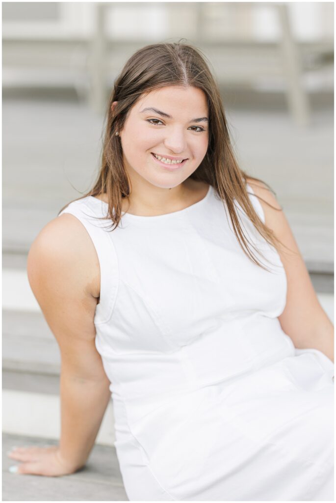 Close-up of the girl seated on the steps, wearing a white dress. She looks directly at the camera with a relaxed smile. Her hair is parted in the middle and falls over her shoulders.