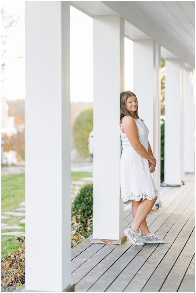 The girl stands on a porch, leaning against a white pillar. She is dressed in a sleeveless white dress and silver sneakers, looking towards the camera with a gentle smile. The porch overlooks a green lawn with distant greenery in soft focus.