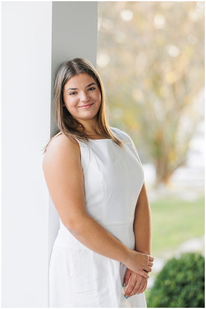 Close-up of the senior girl leaning against a white pillar outdoors. She wears a sleeveless white dress and smiles softly at the camera. The background shows hints of greenery with a soft, warm light.