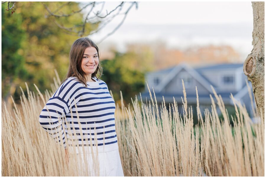 The girl stands in tall, tan grass outdoors, wearing a navy and white striped sweater over a white skirt. She smiles as she looks toward the camera. In the background, a house with gray siding and a gable roof is partially visible, surrounded by trees.