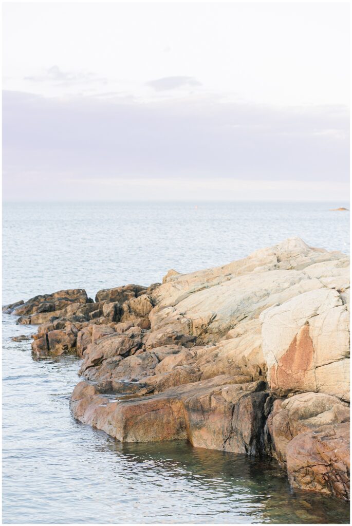 Coastal scene in Cohasset, MA with large, rugged rocks along the water’s edge. The ocean is calm, and the sky is pale, with soft clouds stretching across the horizon.