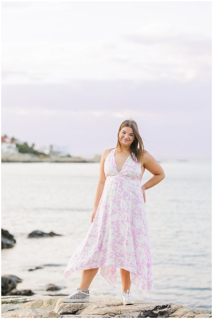 The girl stands on rocks by the ocean, wearing a flowing white and pink floral dress with a deep v-neckline and silver sneakers. She smiles gently, one hand on her hip. The water and a few distant houses are visible behind her under a cloudy sky.