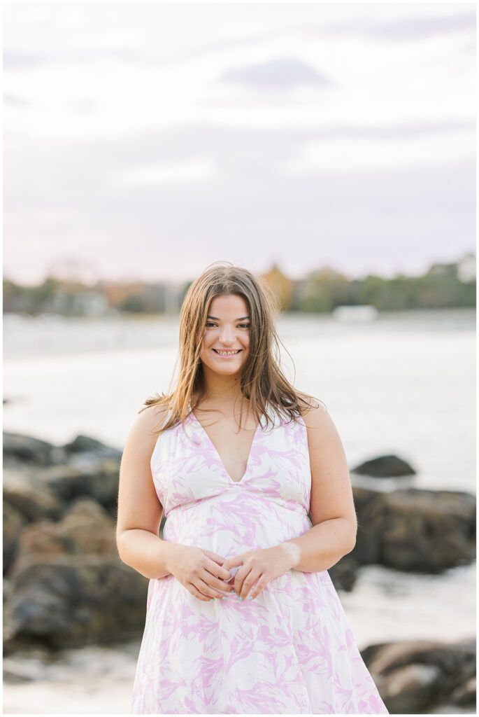 The girl stands near the shoreline, wearing a white and pink floral dress, smiling with her hands clasped in front of her. The background features rocky terrain and calm ocean water under a cloudy sky.