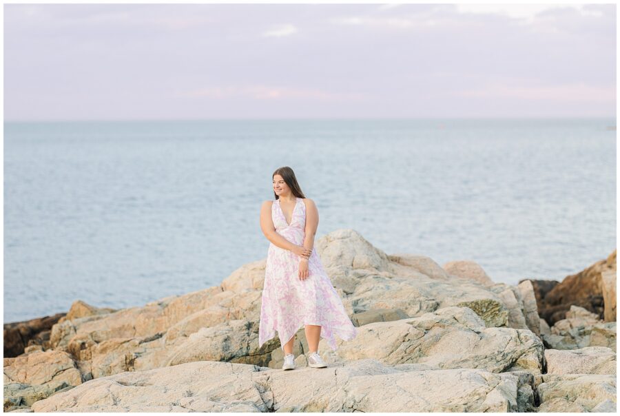 A high school senior girl stands on large coastal rocks with the ocean in the background. She wears a flowing white and pink floral dress and silver sneakers, looking off to the side with a soft smile. This image highlights the Cohasset Beach setting.