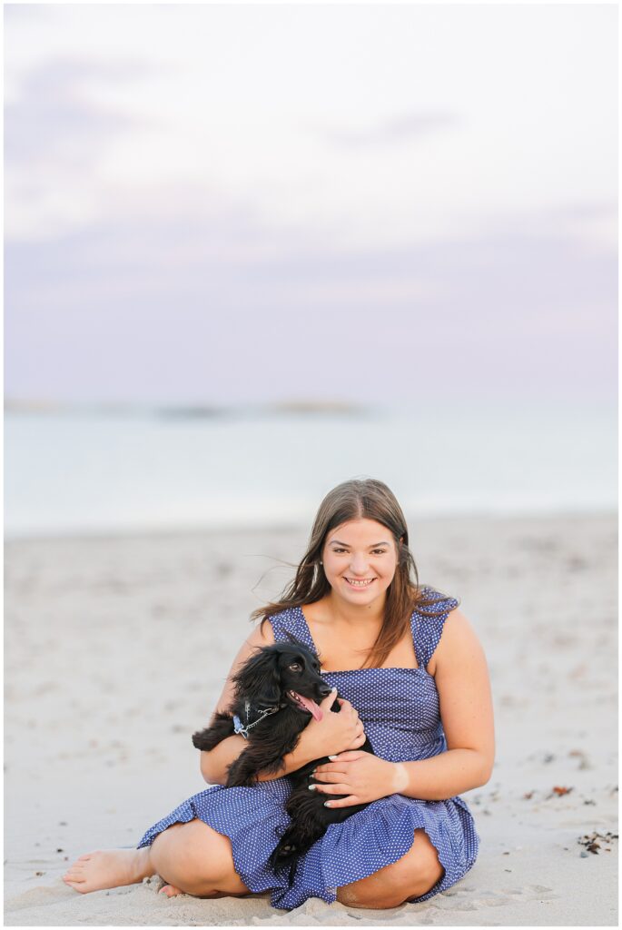 The senior girl sits barefoot on the beach, smiling as she holds a small black dog in her lap. She is dressed in a blue polka-dot sundress, and the calm ocean stretches out behind her. This beach scene is captured at Sandy Beach in Cohasset.