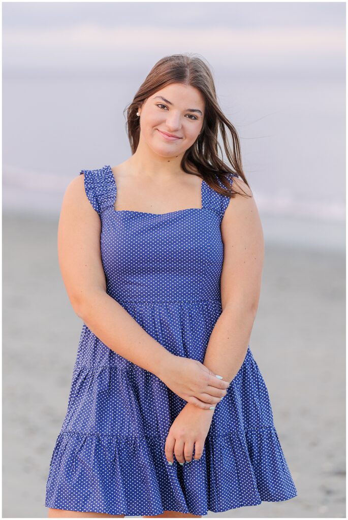 The senior girl stands on the beach in a blue polka-dot sundress, smiling with her hands clasped in front of her. The soft, pastel tones of the ocean and sky create a calm atmosphere at Sandy Beach in Cohasset, MA.