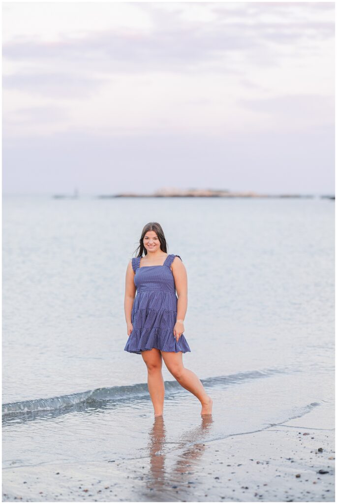 The senior girl stands ankle-deep in gentle ocean waves at Sandy Beach in Cohasset, MA., wearing a blue polka-dot dress. She smiles towards the camera, with the distant ocean horizon visible behind her.