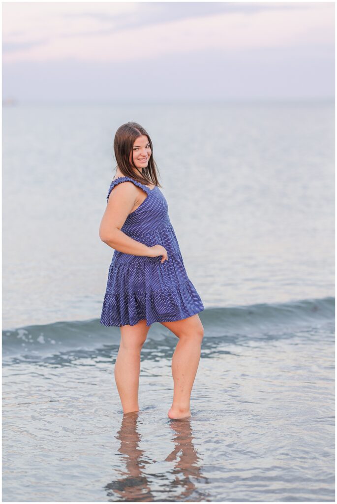 Side view of the girl standing barefoot in shallow water at Sandy Beach in Cohasset, MA. She smiles and glances back over her shoulder, wearing a blue polka-dot sundress, with calm waves and an expansive ocean view in the background.