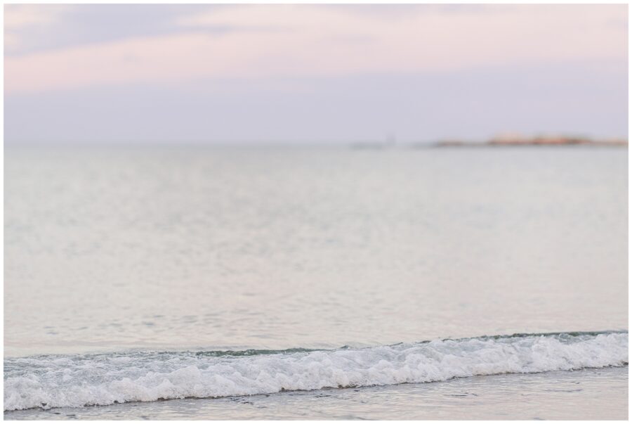 Close-up of small, gentle waves lapping at the sandy shore at Sandy Beach in Cohasset, MA. The water is calm, and the sky above is soft and pastel, enhancing the peaceful beach scene.