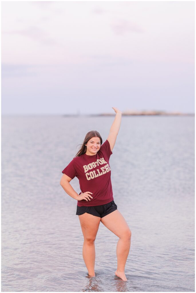 High school senior girl standing ankle-deep in the water at Sandy Beach in Cohasset, MA, with a pastel sky in the background. She wears a maroon Boston College t-shirt and black shorts, smiling and raising one arm playfully. This senior portrait captures a fun moment with the Cohasset Beach backdrop.