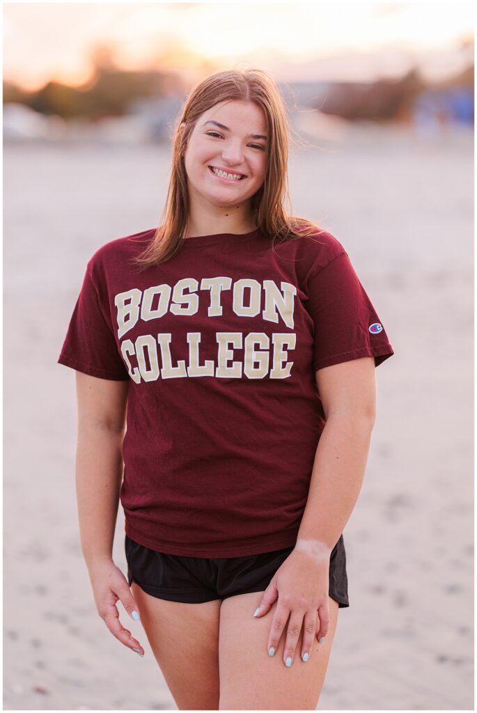 Close-up of the senior girl on Sandy Beach in Cohasset, MA, smiling warmly at the camera. She is wearing a maroon Boston College t-shirt and black shorts, with a softly lit, sandy beach scene in the background. Perfect for a Cohasset Beach Photographer setting.