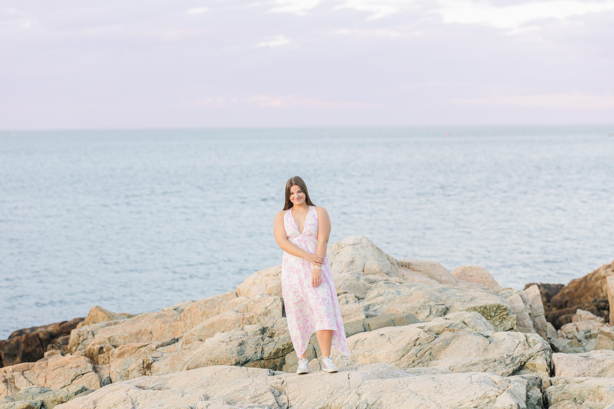 High school senior girl standing on large rocks by the ocean at Sandy Beach in Cohasset, MA. She wears a white and pink floral dress, looking directly at the camera with a gentle smile. This portrait showcases the scenic Cohasset Beach backdrop for a senior photoshoot.