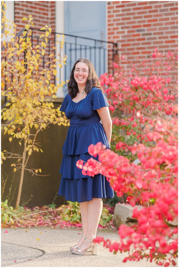 A high school senior stands smiling outdoors, surrounded by bright red bushes and yellow fall foliage. She wears a layered navy dress and silver heels, posing in front of a brick building in Lynn, MA.