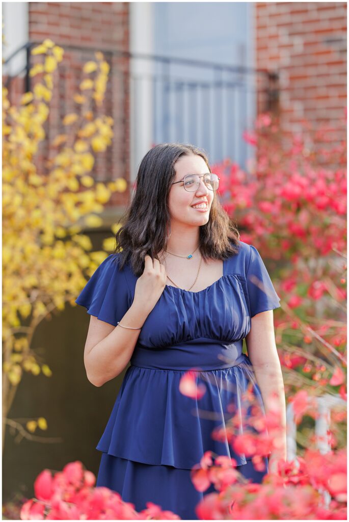 A senior girl with glasses and dark, shoulder-length hair smiles while standing near a red bush. She is wearing a navy blue dress, with a yellow tree and brick building in the background, perfect for fall senior pictures.