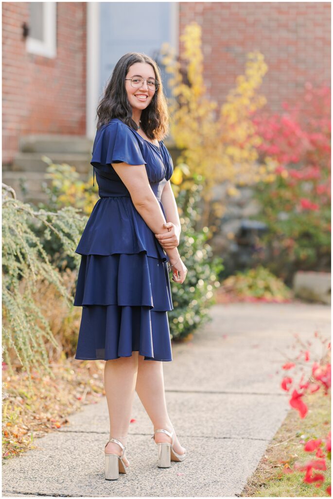 A smiling senior poses on a paved path near a brick building with red bushes and yellow trees. She wears a navy layered dress, silver heels, and glasses, ideal for showcasing fall senior pictures.