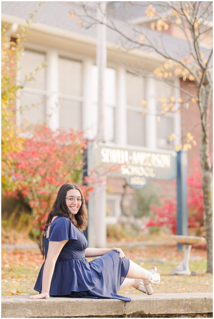 A high school senior sits on a ledge in front of a sign reading “Sewell-Anderson School, 1920.” She wears a layered navy dress and smiles, surrounded by colorful autumn trees in Lynn, MA.