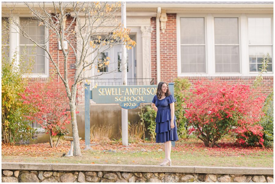 A young woman stands near a large “Breed Middle School” sign, surrounded by green shrubs and fall foliage. She wears a navy blue dress and smiles brightly for her senior pictures.