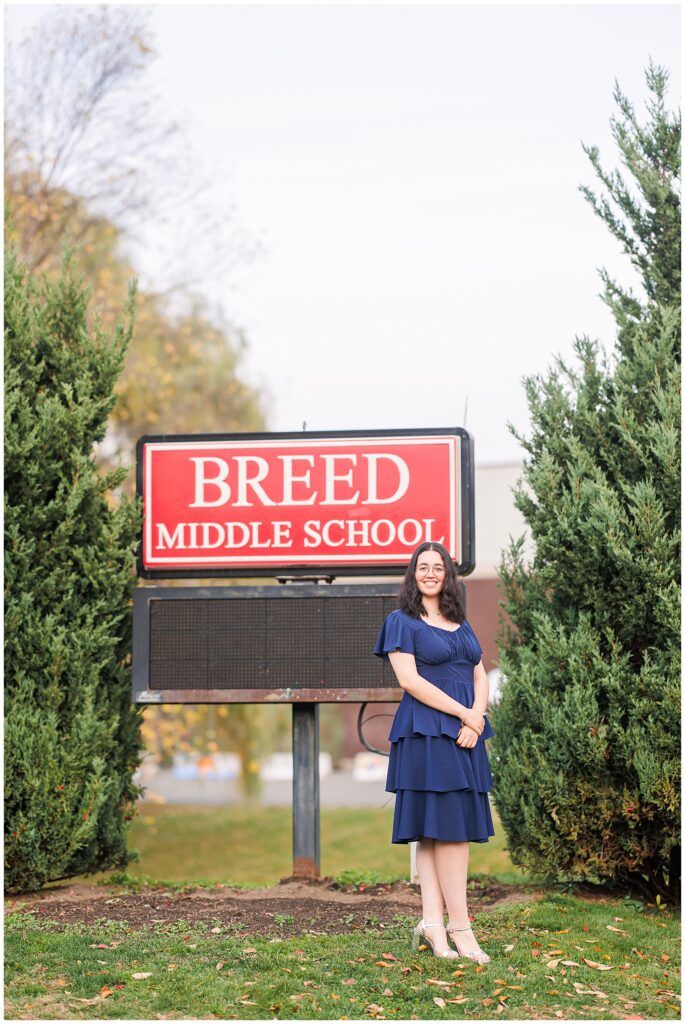 A senior walks on a sidewalk with a large, vibrant orange tree in the background. She is wearing a navy blue dress and silver heels, capturing the beauty of fall senior pictures in Lynn, MA.