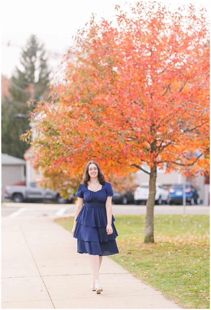 A senior walks on a sidewalk with a large, vibrant orange tree in the background. She is wearing a navy blue dress and silver heels, capturing the beauty of fall senior pictures in Lynn, MA.