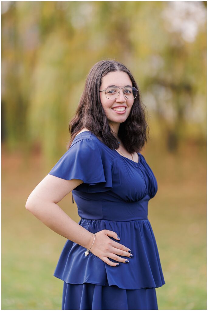 A close-up of a smiling senior standing outdoors in front of blurred greenery. She wears glasses, a navy blue dress, and a simple bracelet, highlighting her natural look during fall in Lynn, MA.