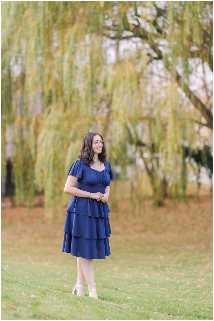 A senior girl stands in a grassy park with cascading willow trees in the background. She is wearing a navy layered dress and silver heels, posing naturally during fall in Lynn, MA.