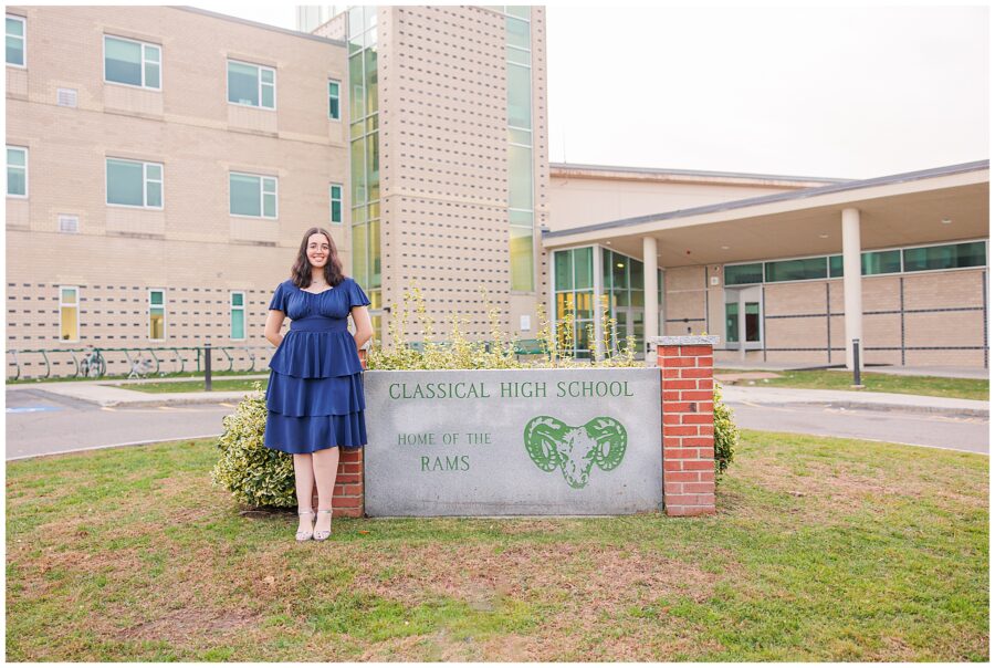 A high school senior poses next to a sign that reads “Classical High School, Home of the Rams.” The beige school building is in the background, and she is wearing a navy blue dress and smiling brightly.