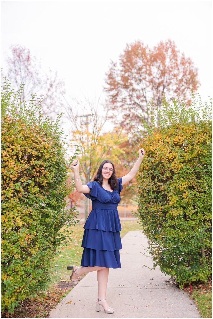 A young woman smiles and raises her arms in a playful pose between two trimmed hedges. She is wearing a navy blue dress with fall trees featuring orange and yellow leaves visible in the background, perfect for fall senior pictures in Lynn, MA.