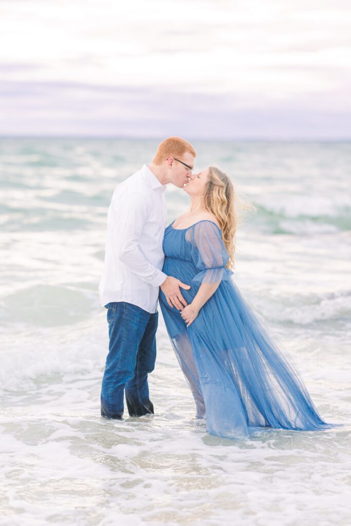 A couple stands in the shallow water at the beach during daytime, sharing a kiss. The man wears a white shirt and dark jeans, and the woman, visibly pregnant, wears a flowing blue sheer dress. Gentle waves surround them under a cloudy sky. Falmouth, MA Cape Cod