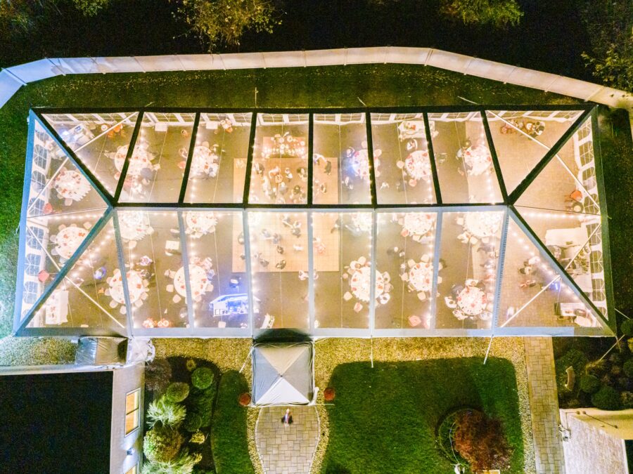 An aerial nighttime view of a clear tent set up for an event. Inside, round tables with white tablecloths and chairs are illuminated by string lights, and guests can be seen mingling.