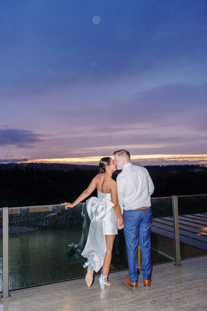 A couple shares a kiss on a balcony at sunset, overlooking a scenic horizon. The woman wears a white satin dress with a large bow on the back, and the man wears a white shirt and blue trousers. The sky is a mix of purple, blue, and pink hues. New Hampshire wedding photographer