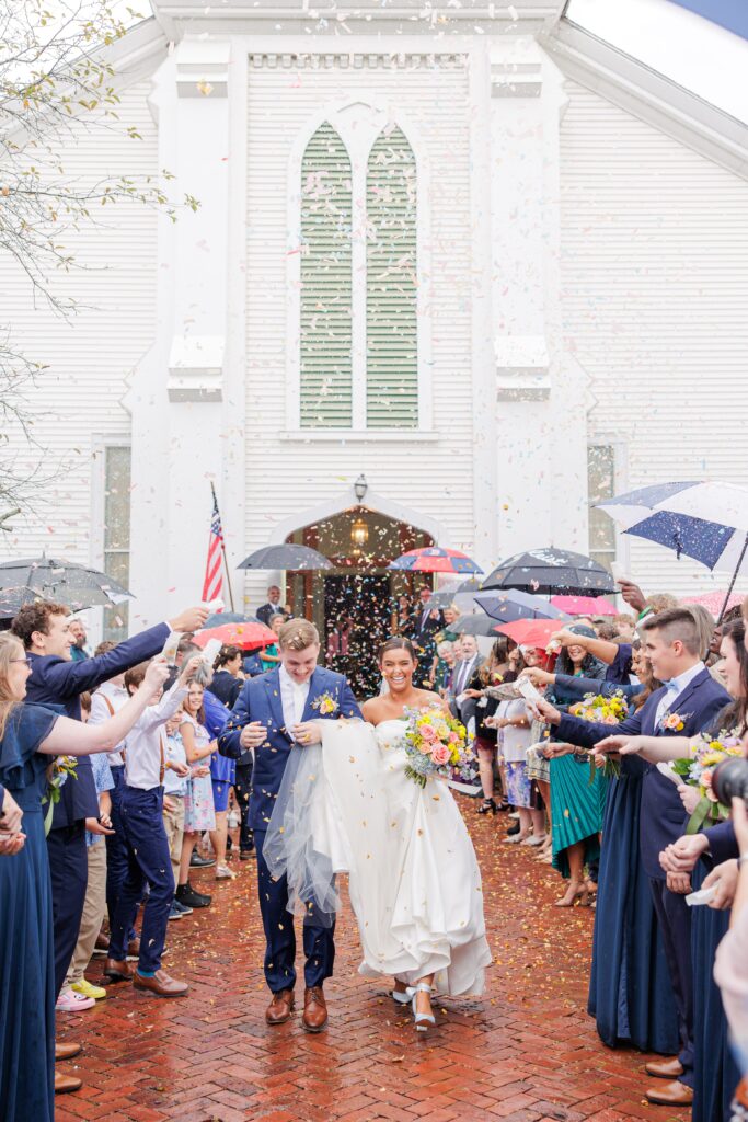 A newlywed couple walks out of a white church as guests shower them with colorful confetti. The groom wears a navy suit, and the bride wears a white wedding dress while holding a bouquet of vibrant flowers. Guests hold umbrellas in the background. New Hampshire wedding photographer