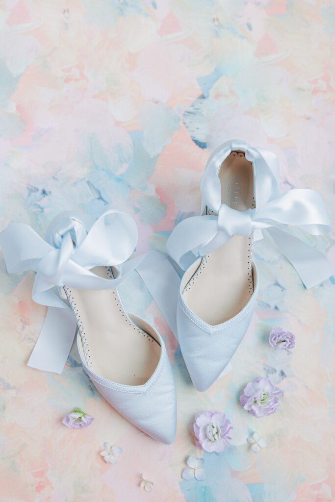 A pair of blue bridal shoes with large satin bows are displayed on a pastel floral background, surrounded by small flowers in shades of purple and white.