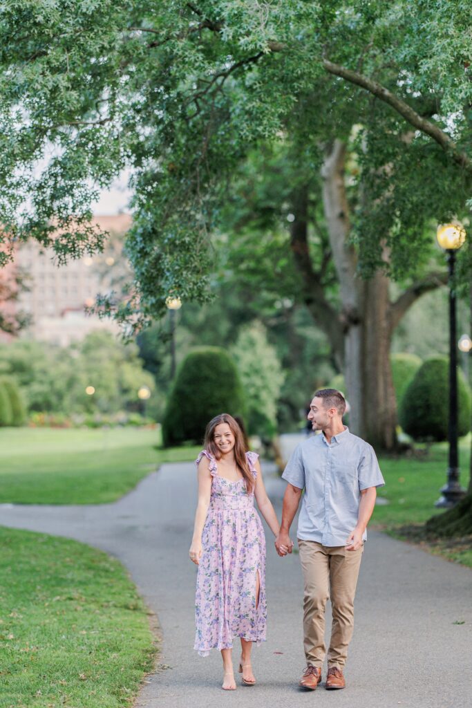 A smiling couple walks hand-in-hand on a park path lined with green trees. The woman wears a floral pink dress, and the man wears a light blue shirt and beige pants. Lampposts and park greenery line the walkway. Boston Public Garden