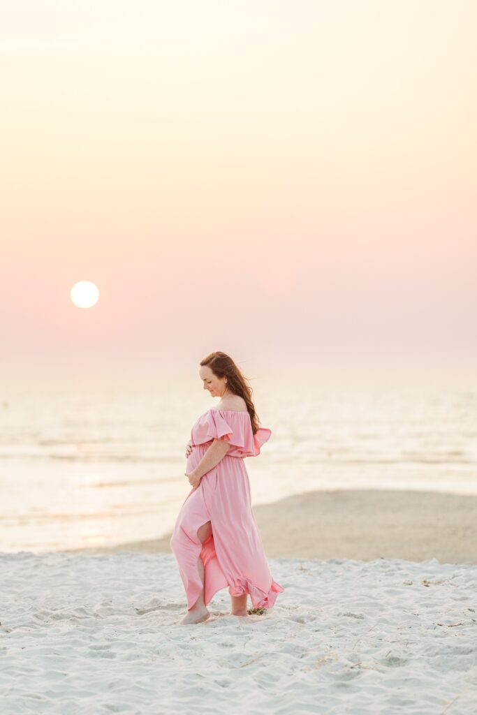 A pregnant woman in a flowing pink dress stands on a sandy beach at sunset. She cradles her belly and looks down gently as the sun glows in the sky, casting soft light over the calm sea. Mayflower Beach Cape Cod